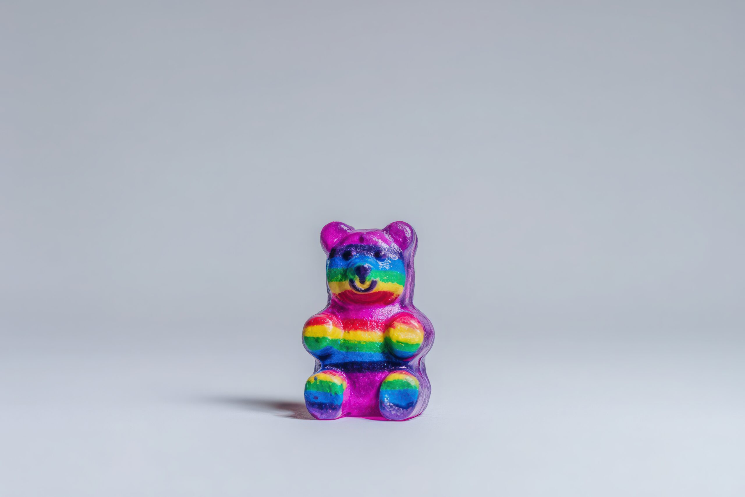 A vibrant gummy bear candy features a rainbow striped pattern, showcasing bright colors. The bear sits proudly on a plain surface, creating a cheerful focal point.