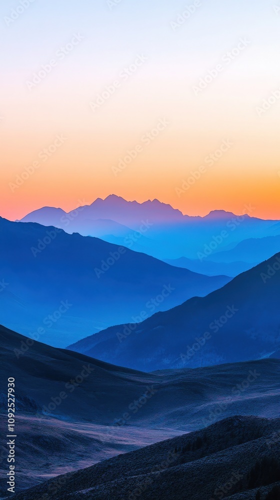 A stunning view of mountains at dawn showcases layers of peaks bathed in orange and blue hues. The soft light creates a tranquil atmosphere, highlighting the natural beauty of the landscape.