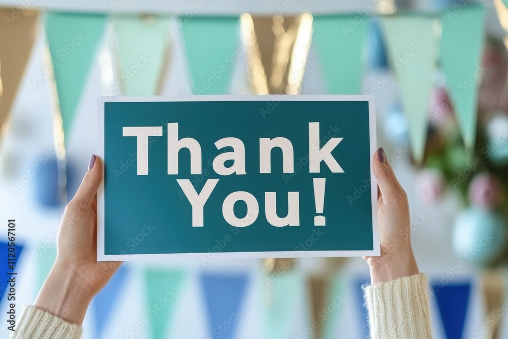 Two hands are raised, displaying a blue sign with bold white letters stating thank you. The background features colorful decorations, creating a cheerful and festive atmosphere for a celebration.