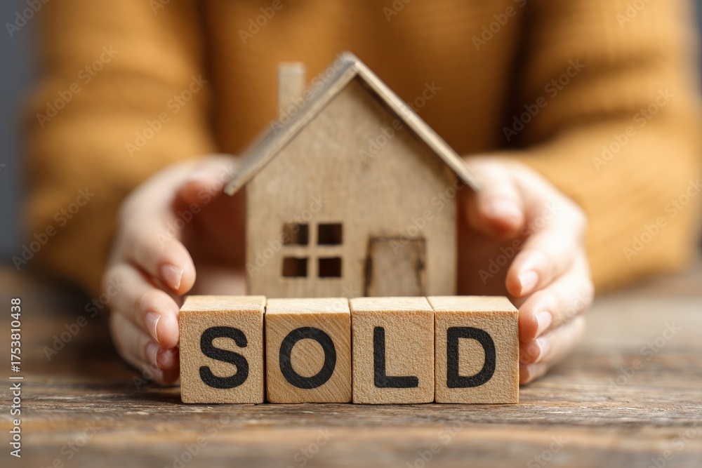 A person is holding a small house model with blocks that spell sold. This scene conveys the excitement of closing a real estate deal. The setting is warm and inviting, suggesting a cozy atmosphere.